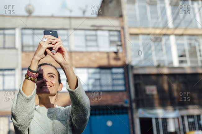 Mid aged woman taking a selfie with a mobile device in urban scenery at sunset with half face in shadow