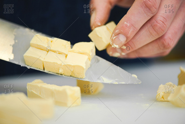 Close-up view of human hand cutting butter into cubes with big steel kitchen knife on white chopping board
