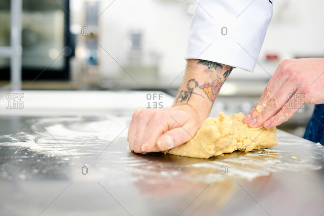 Making cookie dough. Close-up view of male hands with tattoo kneading dough on steel table in restaurant kitchen