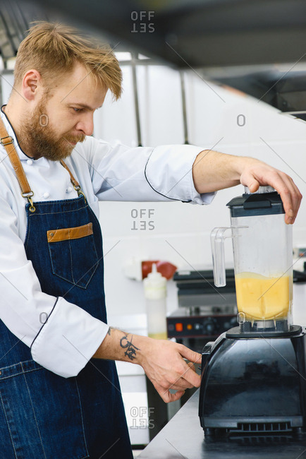 Working in restaurant. Blond bearded cook in denim apron making delicious orange smoothie in modern blender