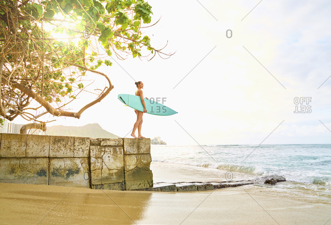 Pacific Islander woman holding surfboard on wall near ocean