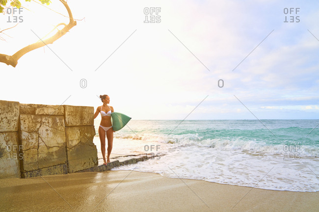 Pacific Islander woman holding surfboard near wall at ocean