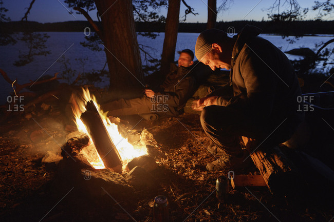 Sweden, Skane, Filkesjon, Mid adult men sitting around campfire at dusk ...