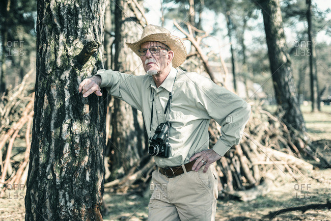 Senior male nature photographer wearing hat leaning against tree.