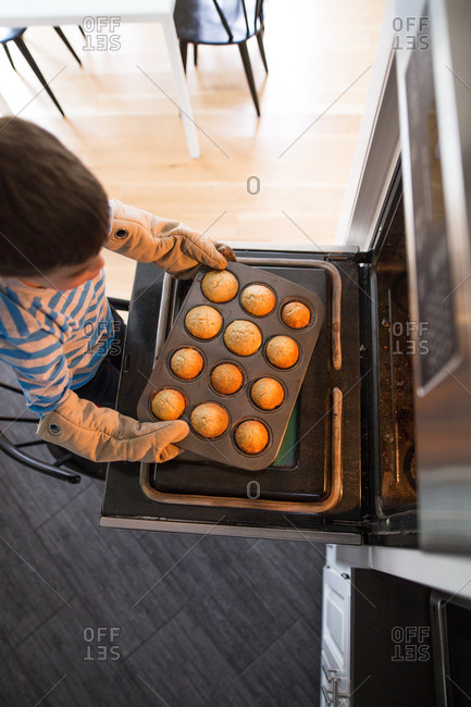Boy wearing oven mitts taking pan with freshly baked muffins out of the oven