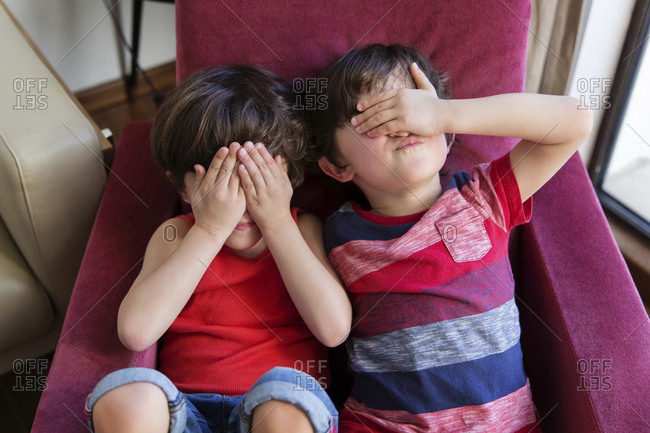 Twin brothers sitting together on arm chair covering eyes with hands
