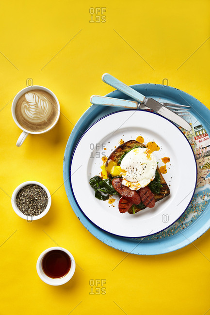 Poached egg on toast with spinach, fried chorizo and paprika oil on yellow surface and large round graphic serving tray.