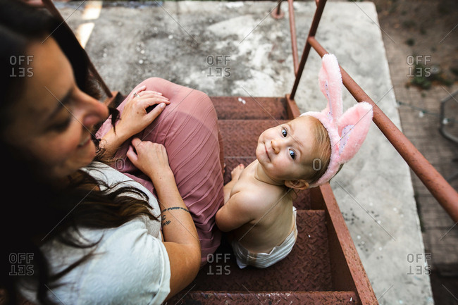 Mom and diapered baby sit outside on fire escape while wearing bunny ears