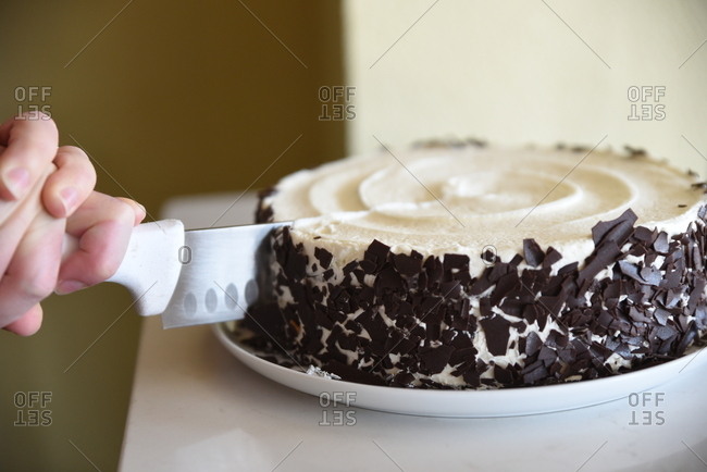 Person slicing a cake with vanilla icing and chocolate shavings
