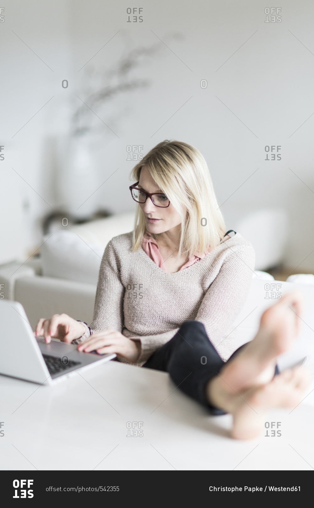 Blond woman sitting at table with feet up using laptop stock photo - OFFSET