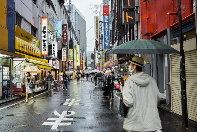 Tokyo, Japan - July 6, 2015: Man wearing facemask observes surrounding electronics stores as local Japanese businesspeople walk by him in the rain