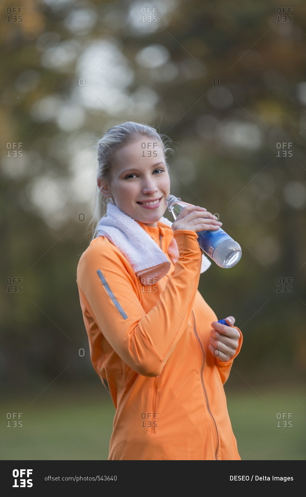 Pretty blonde woman drinking water in park after doing sport and smiling at camera
