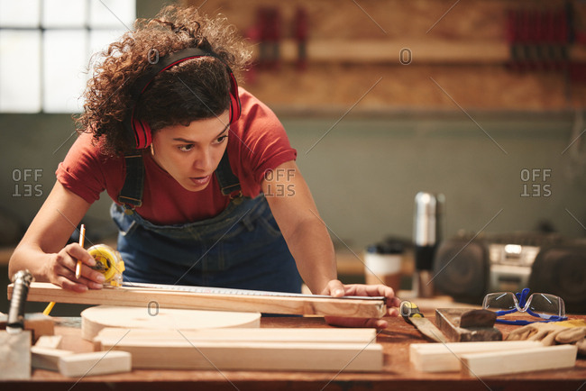 At carpenter workshop. Young concentrated curly woman in ear defenders and overall measuring wooden plank with tape measure