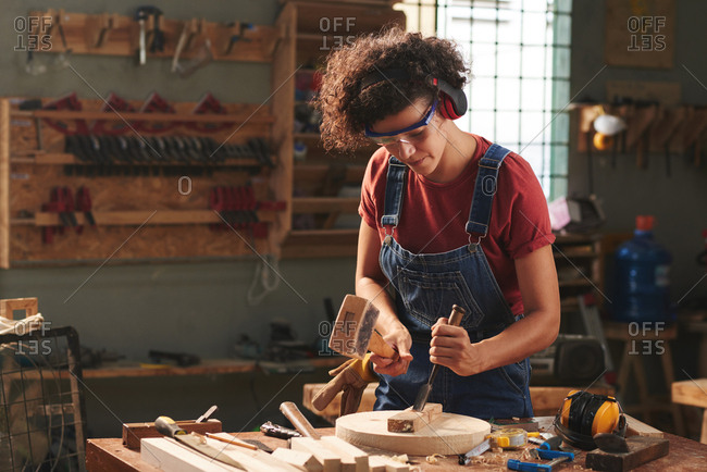 Carpenter in workshop. Concentrated curly woman in ear defenders and goggles using hammer and chisel while working with wooden plank
