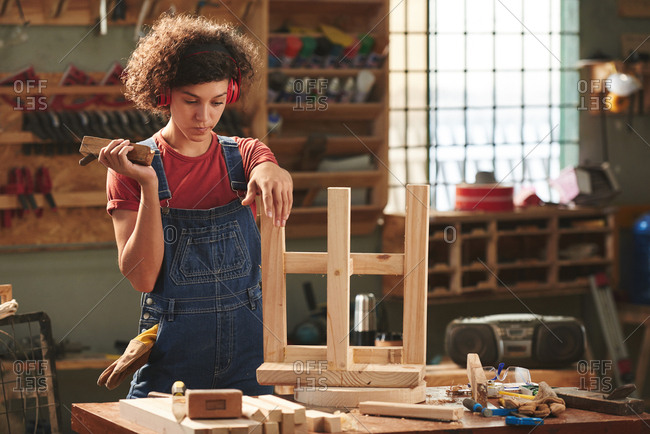 At carpenter workshop. Young curly woman in ear defenders and denim overall holding hand plane and looking at finished wooden stool pensively before polishing it