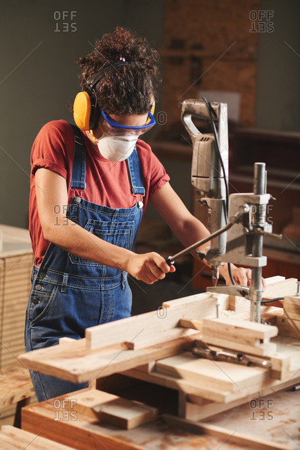 Woodworking industry. Young female carpenter in protective eyewear and ear defenders pressing lever on woodworking machine while cutting wooden planks