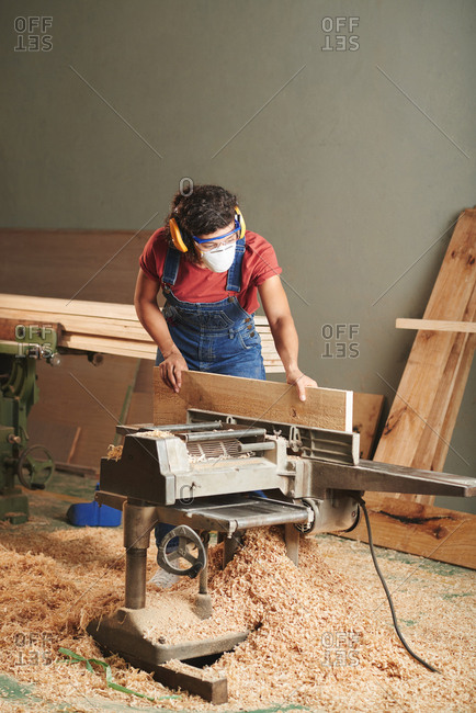Carpentry and joinery. Professional female carpenter in protective eyewear, earmuffs and mask processing wooden plank on woodworking machine. Floor is covered with sawdust