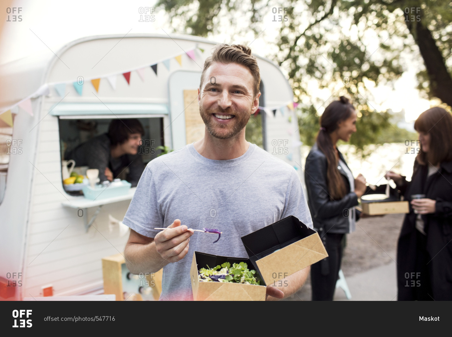 Smiling male customer holding disposable salad box against food truck with friends and owner in background