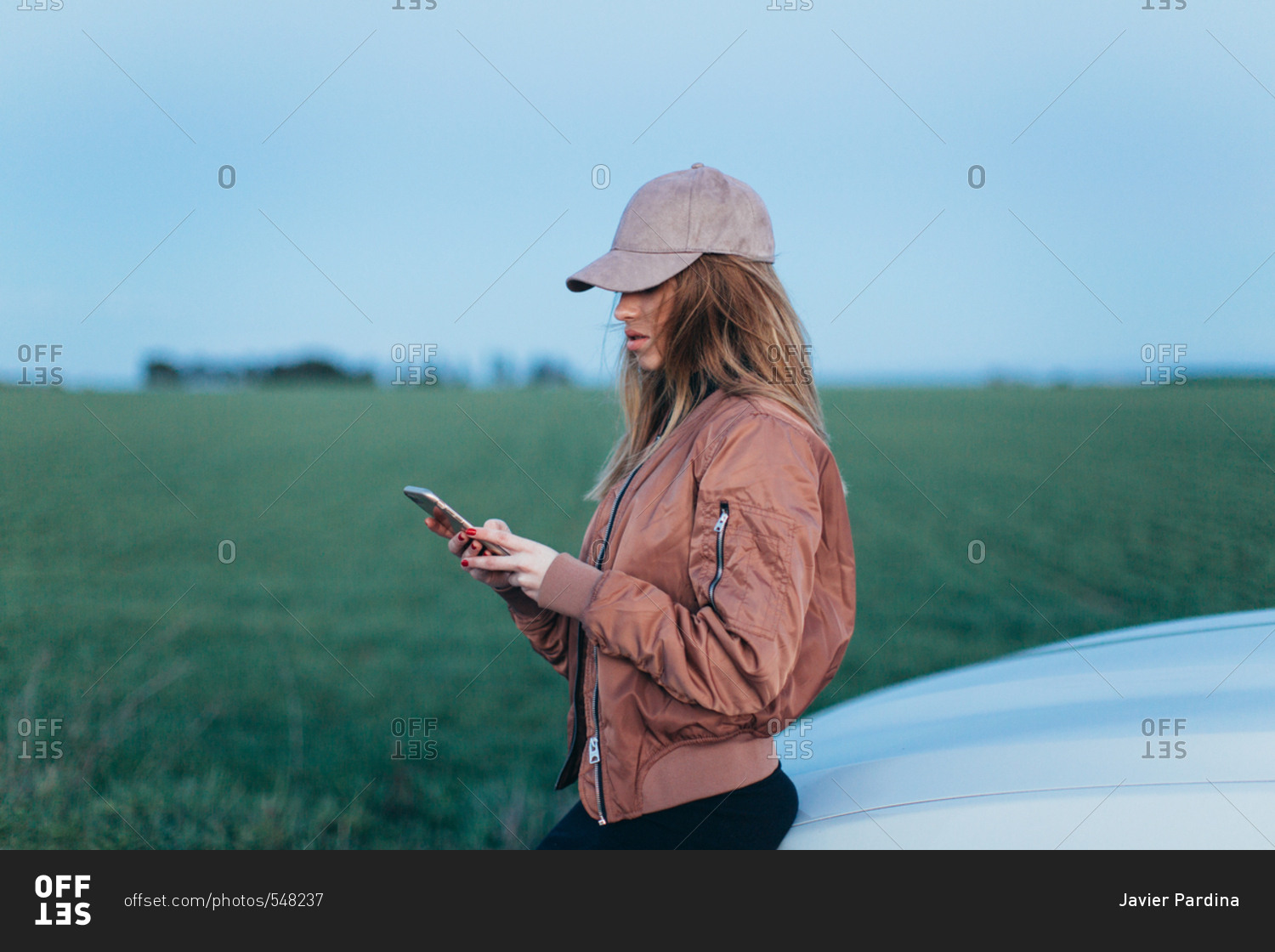 Young woman leaning against a car in the country checking her cellphone