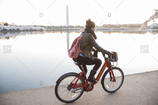 Washington, District Of Columbia, United States - April 12, 2015: A young woman rides a bicycle on the edge of the Tidal Basin in Washington, DC. Blooming cherry blossom trees and the Washington Monument can be seen in the background.