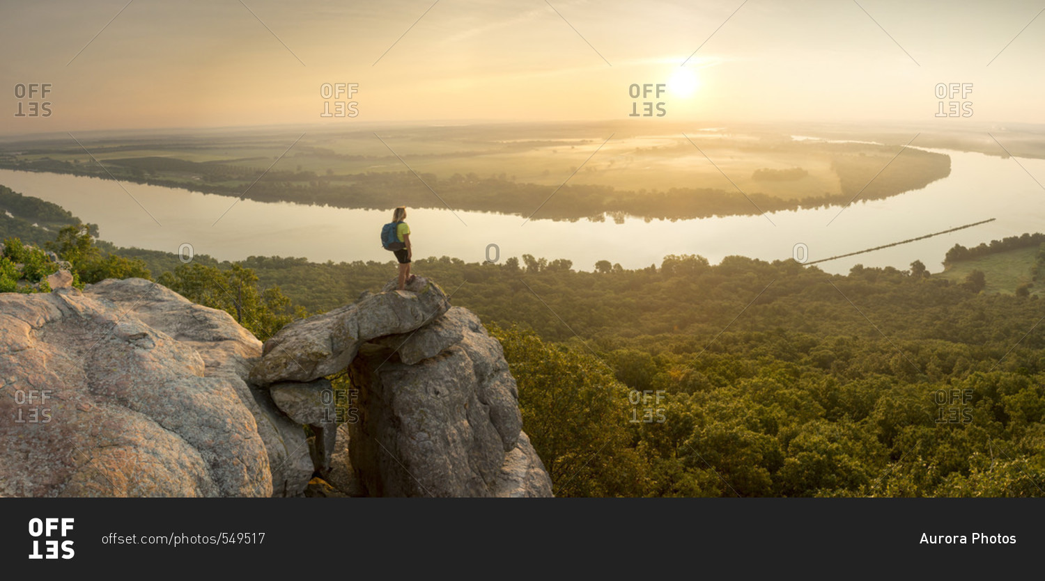 Woman standing on sandstone overhang watching sunrise from summit of Petit Jean Mountain above Arkansas River Valley