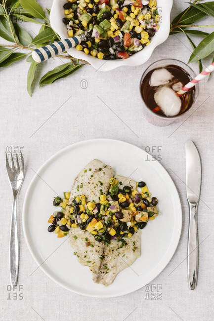 Two plates of Freshly baked tilapia topped off with black bean salsa are photographed on a summery table setting.
