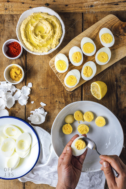 A woman removing yolks from sliced hard-boiled eggs. Sliced eggs, egg whites, hummus and spices accompany.
