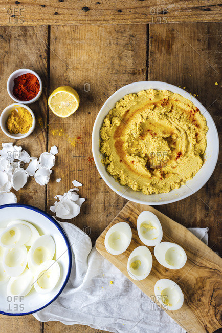 Hummus with hard-boiled egg yolks in a bowl, egg whites in another bowl and on a small wooden cutting board, spices, half lemon and egg shells on a wooden board.