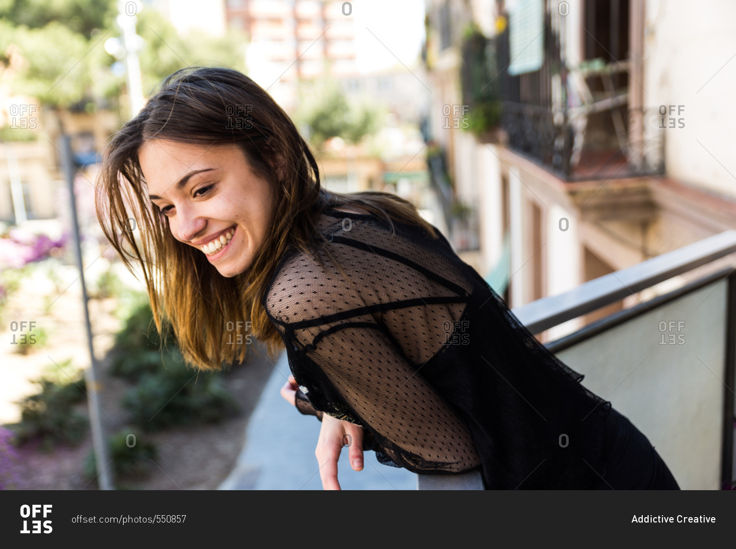 Smiling young girl in black transparent blouse leaning on fence of balcony and looking away.