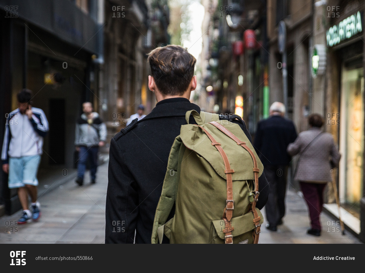 Back view of male in black holding backpack and walking down busy