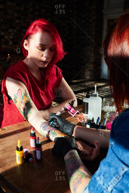 Tattoo lover in studio. Professional tattoo artist using tattoo gun while drawing colorful tattoo on hand of young red-haired woman sitting quietly at table