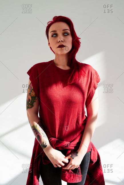 Portrait of young woman with dyed red hair, piercing and light blue eye contact lens standing on white wall background. Her hand covered with colorful tattoos