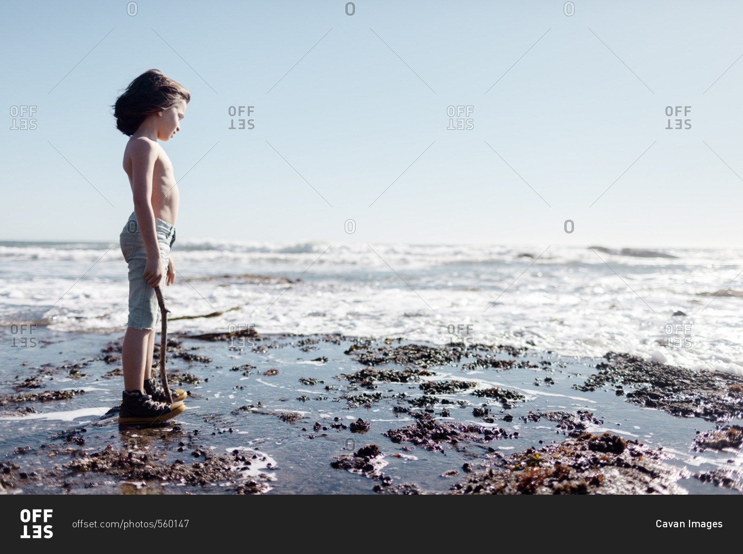 Side view of shirtless boy holding stick standing at beach against clear sky during sunny day