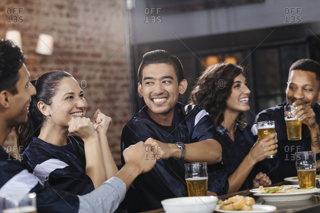 Smiling men giving fist bump while watching sports on TV with friends at bar