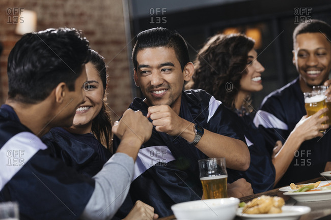 Group of friends giving fist bump while watching sports match with friends at counter in bar
