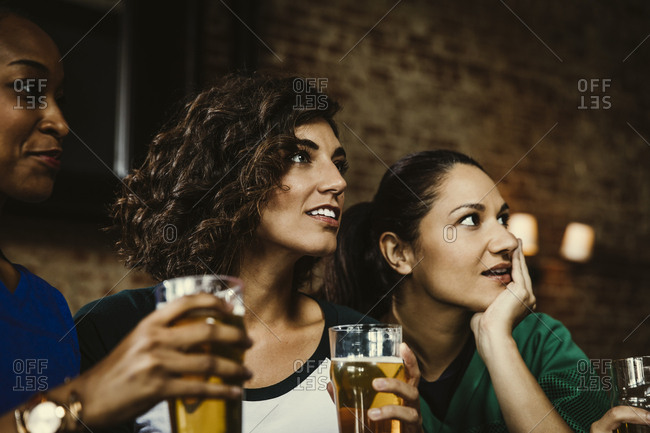 Multiethnic female fans holding beer glasses while watching soccer match in bar