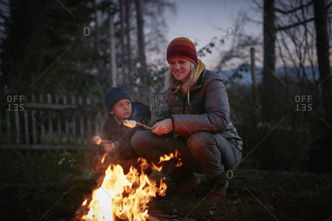 Mature woman and son toasting marshmallows on garden campfire at dusk