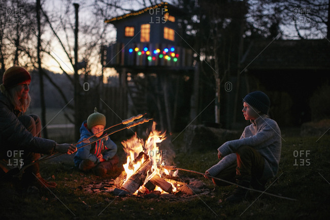 Mature woman and two sons toasting marshmallows on campfire at night