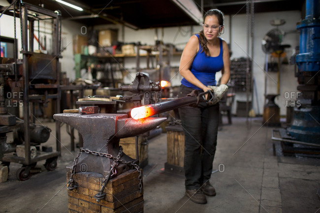 Female metalsmith turning red hot metal rod on workshop anvil