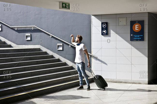 Young man with wheeled trolley bag, in front of steps in train station