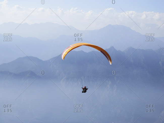 Person skydiving with orange parachute with mountains and clouds in the distance.