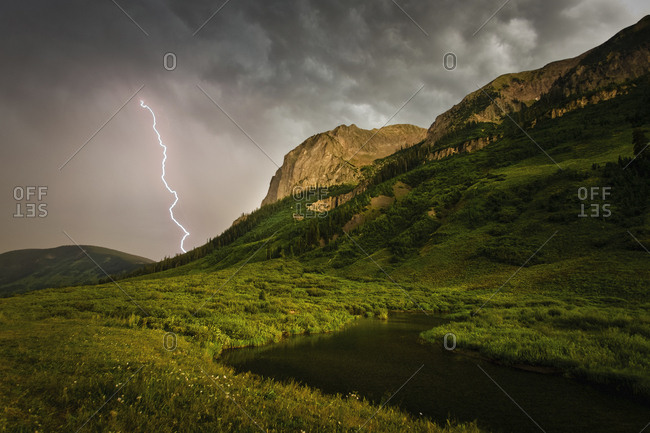 A lightning storm over river running through a mountainous valley. Fork of lightening emerging from clouds.