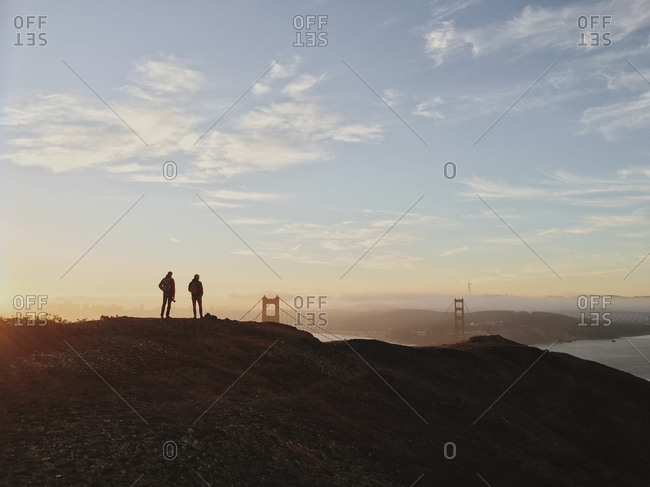 Silhouette of two people standing on a hill overlooking San Francisco Bay, with the Golden Gate Bridge in the distance. Sunset.