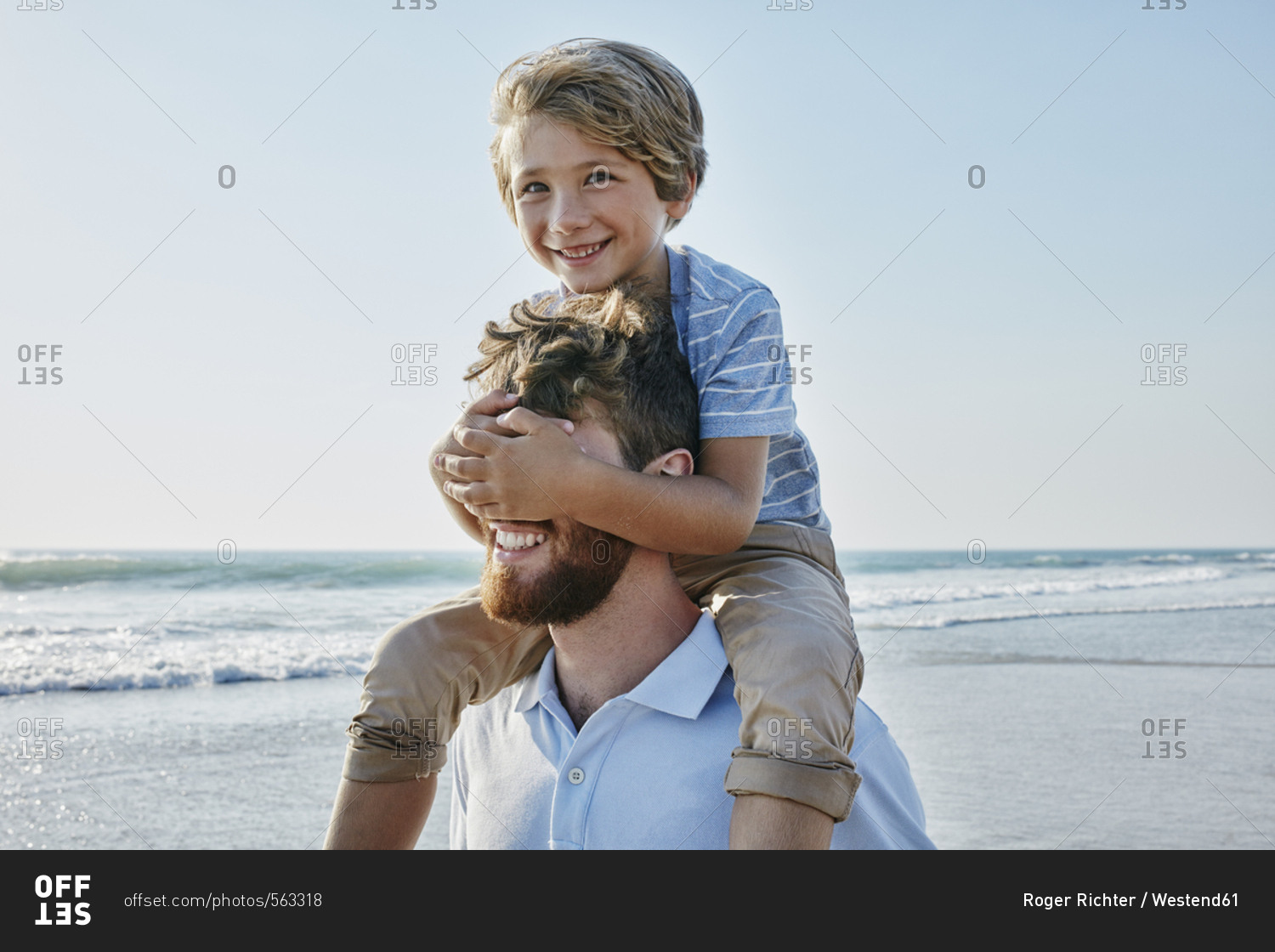 Father carrying son piggyback on the beach- son covering father\'s eyes