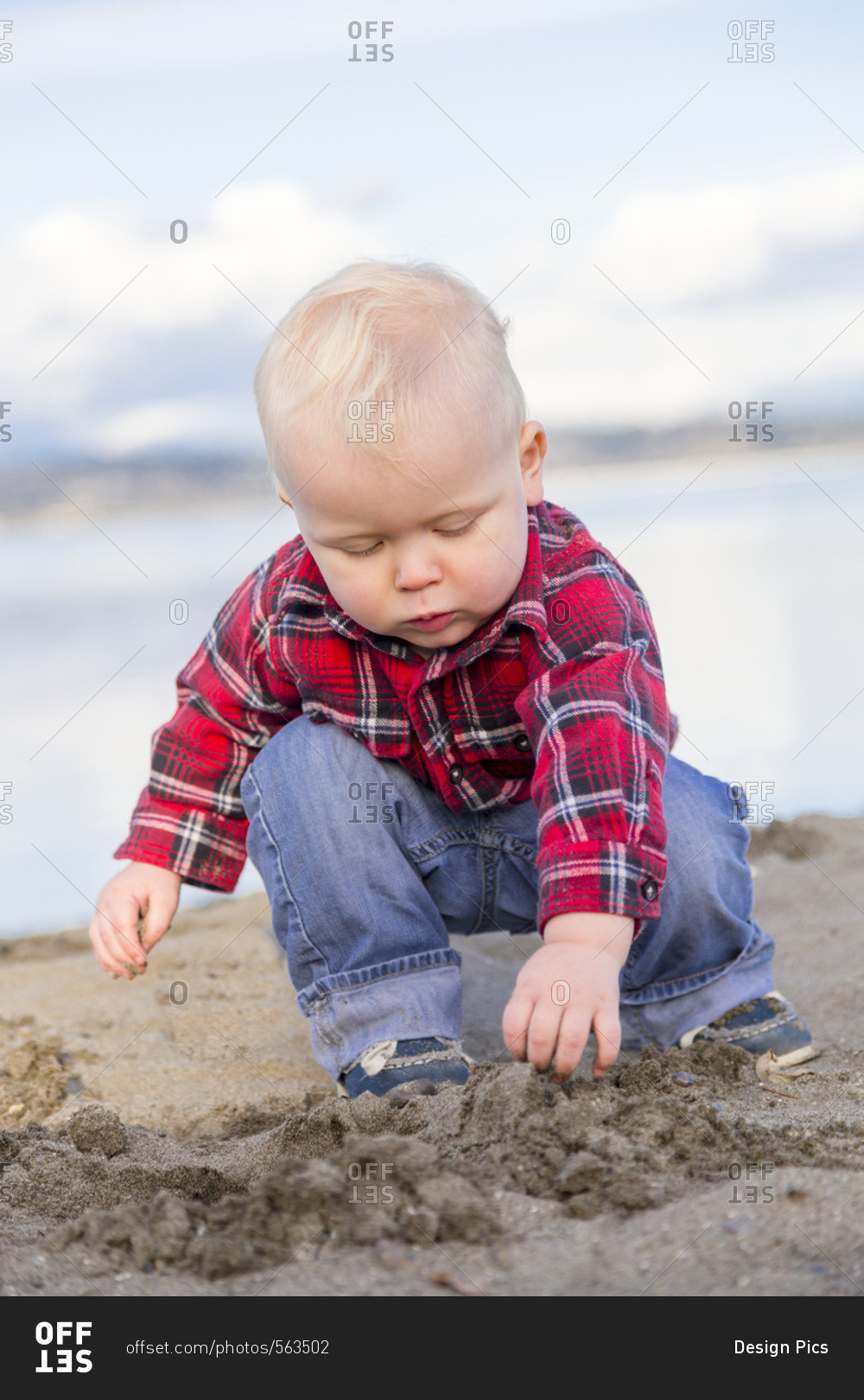 A young boy in a red plaid shirt and jeans plays in the sand by the ocean, Surrey, British Columbia, Canada