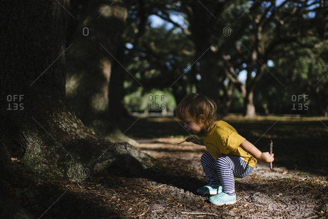 Toddler squatting in a patch of light at a park playing with sticks and dirt
