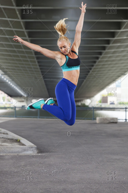 Fit Blonde Woman Jumping In The City Under A Bridge With Arms Raised