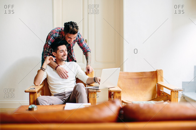 Young man at home, sitting in chair, using laptop, his partner hugging him from behind
