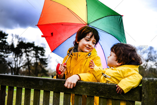 Overhead view of baby boy and brother in yellow anoraks with umbrella on park bench