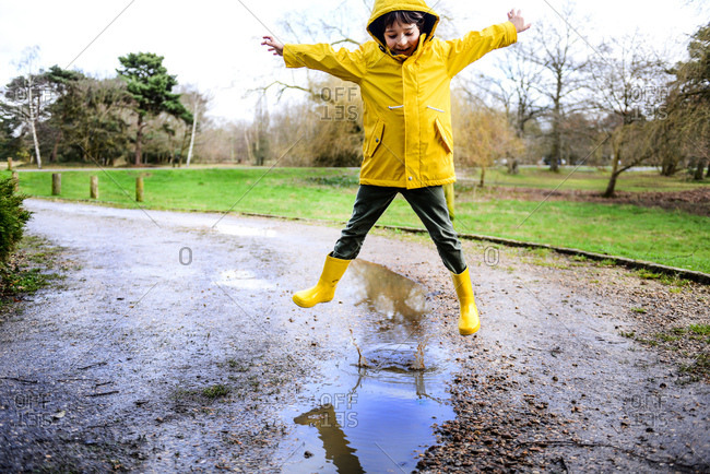 Boy in yellow anorak jumping above puddle in park
